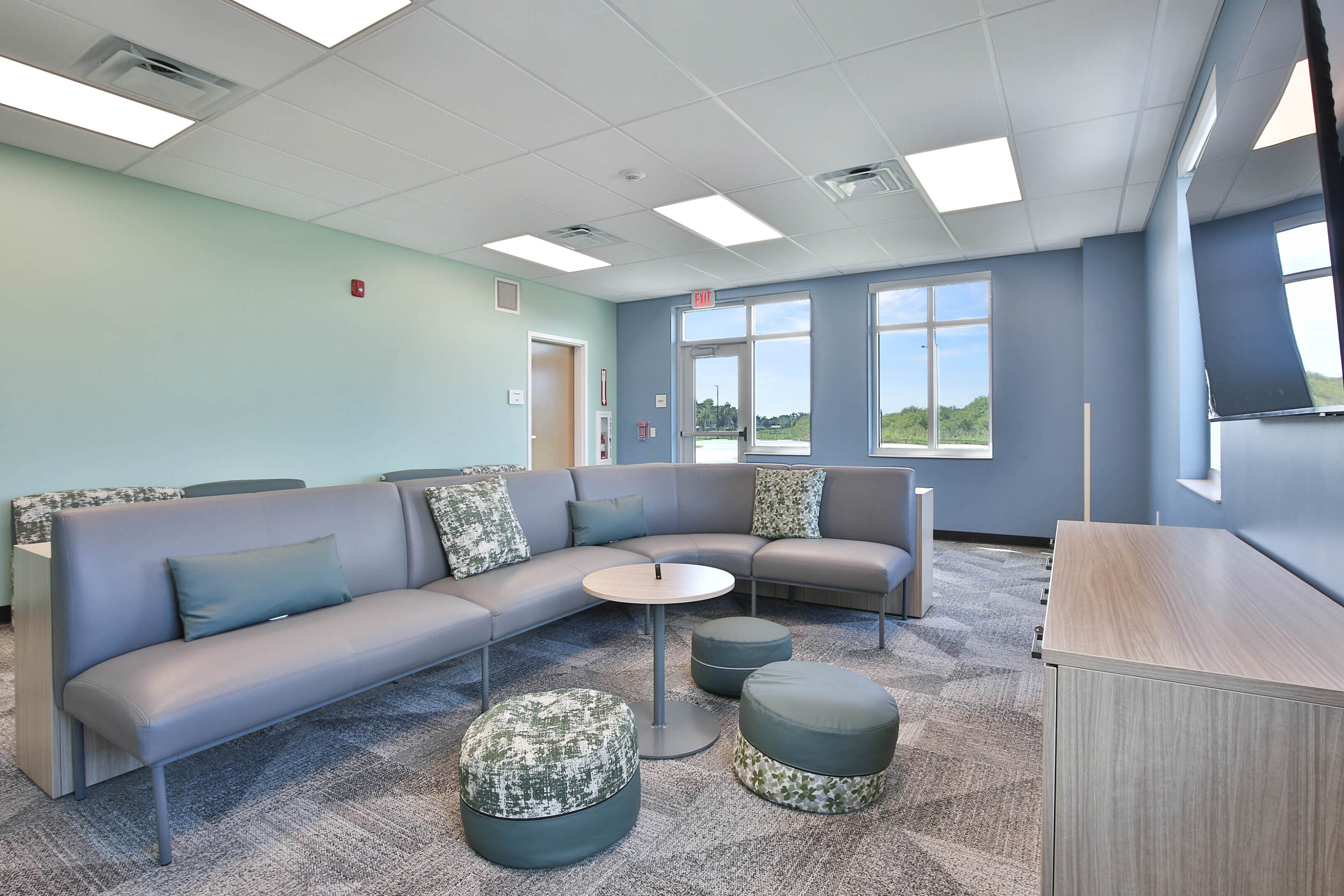 Modern lounge with gray sectional seating, patterned ottomans, a round table, blue accent walls, and gray carpet.