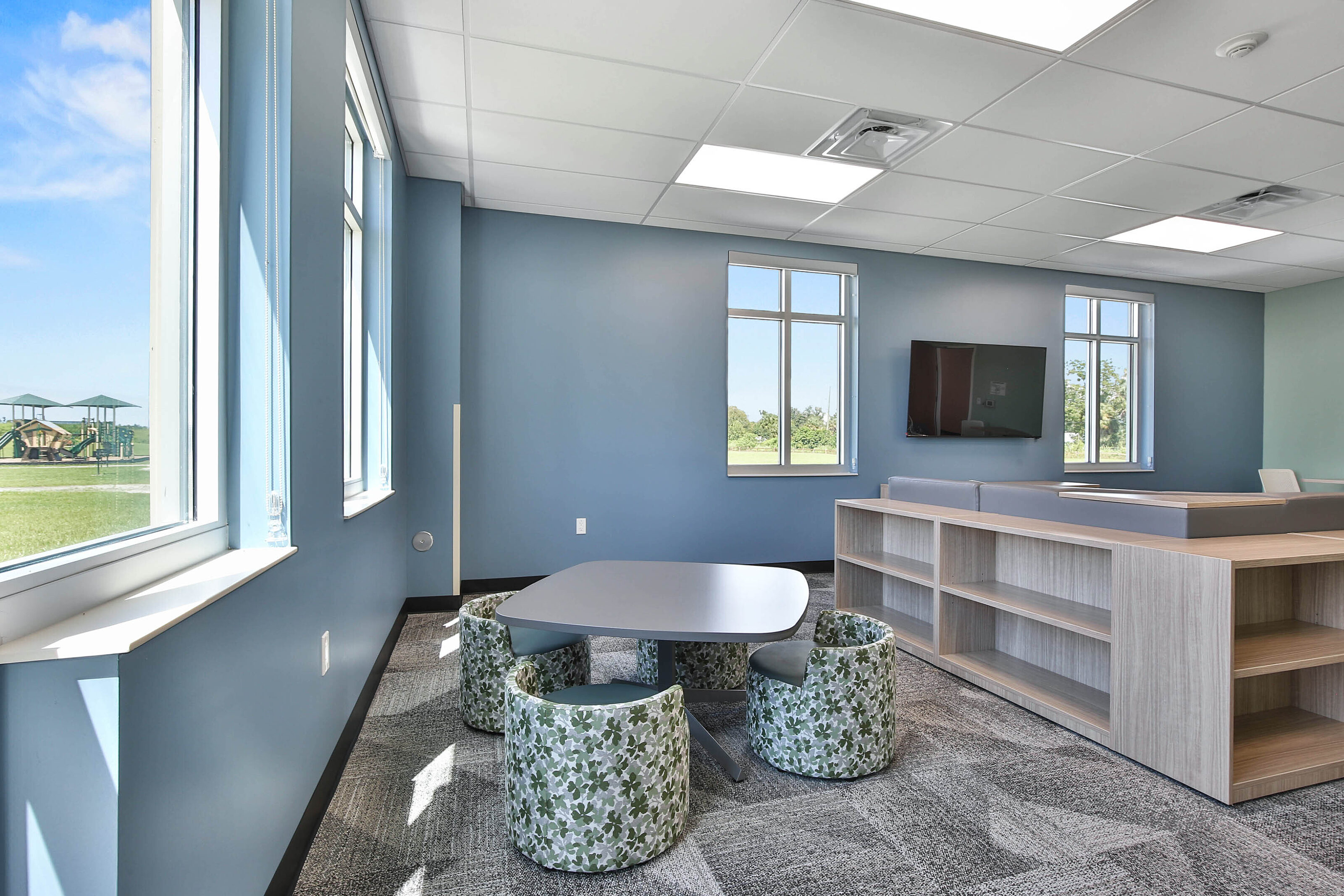Modern lounge with a gray table, green patterned chairs, light wood shelving, and blue walls.