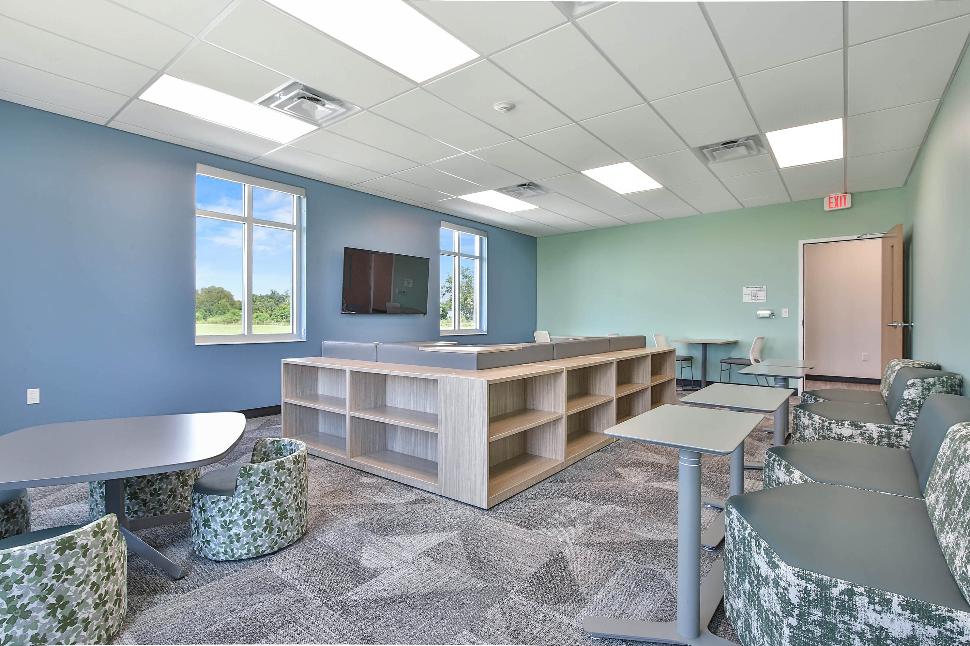 Modern lounge with blue and mint green walls, patterned seating, gray tables, and light wood shelving.