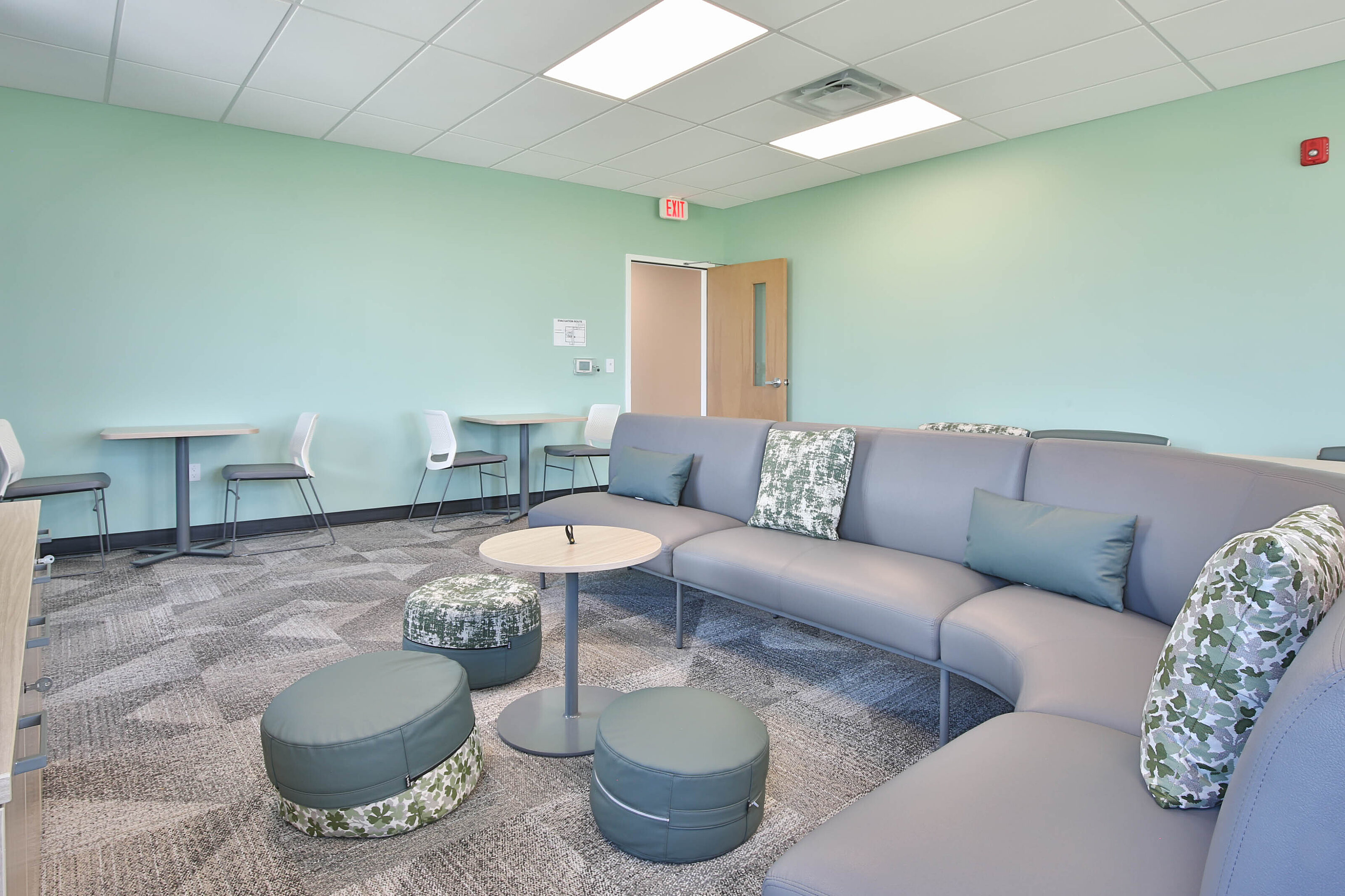 Modern lounge with mint green walls, gray sectional seating, patterned ottomans, and café tables.