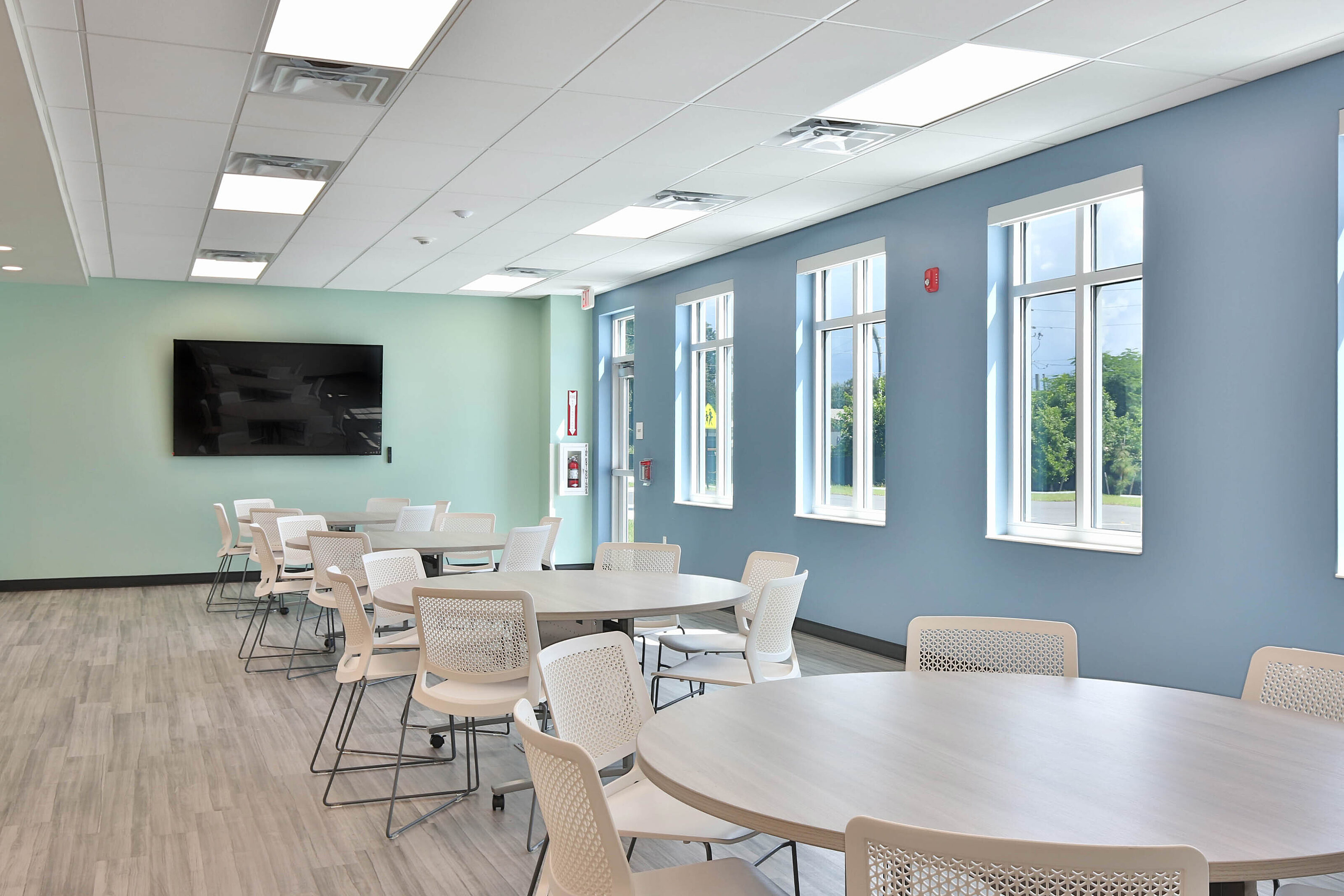 Bright multipurpose room with round tables, white mesh chairs, blue walls, and large windows.