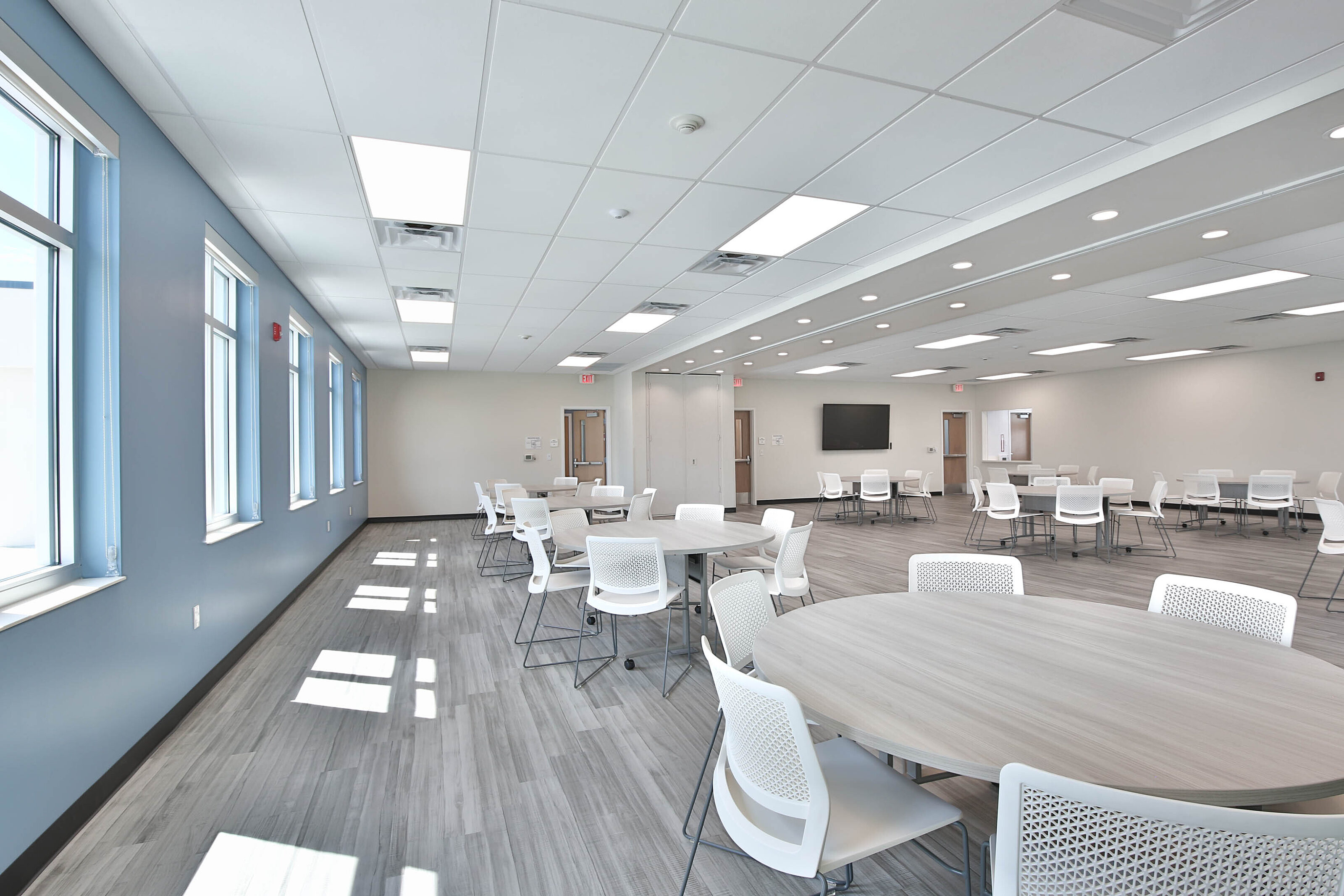 Spacious multipurpose room with round tables, white mesh chairs, blue accent wall, and large windows.
