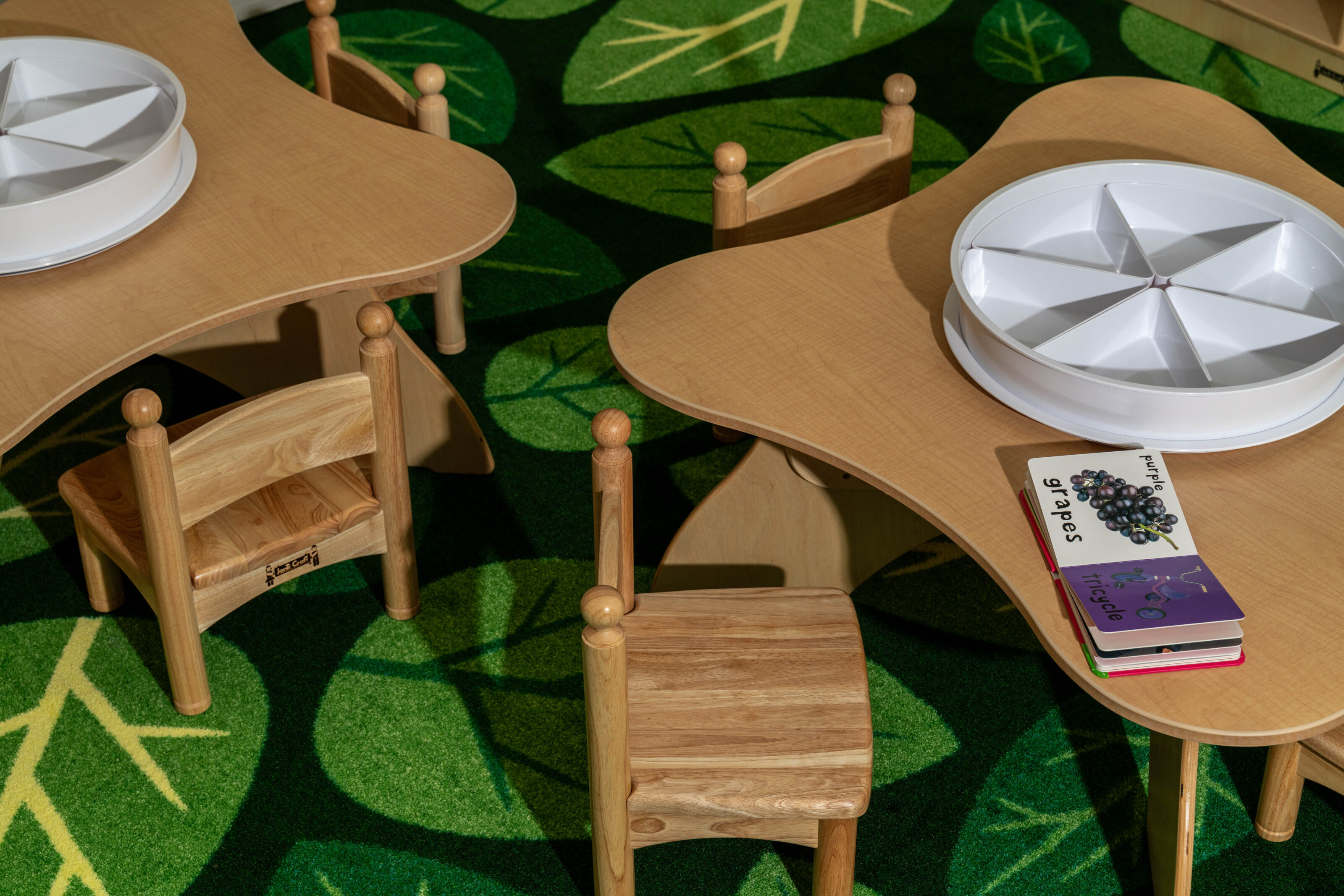 Children’s tables with small wooden chairs, sorting trays, and books on a green leaf-patterned rug.