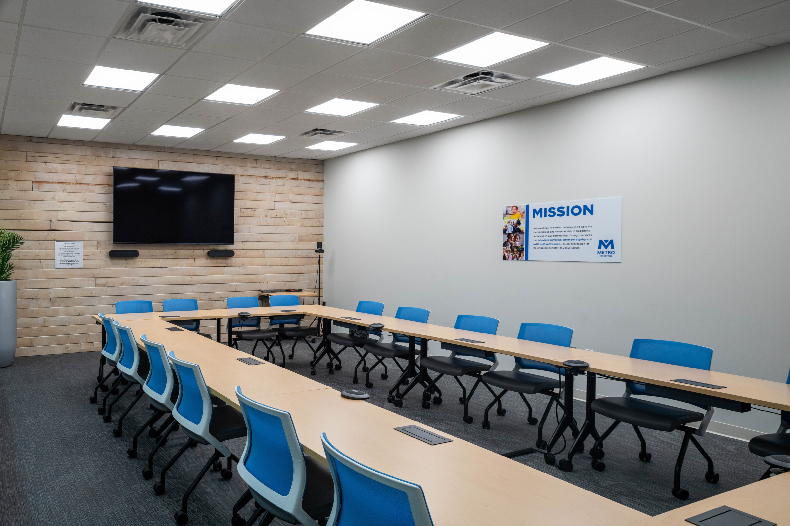 Training room with U-shaped tables, blue chairs, wall-mounted screen, and mission statement poster.