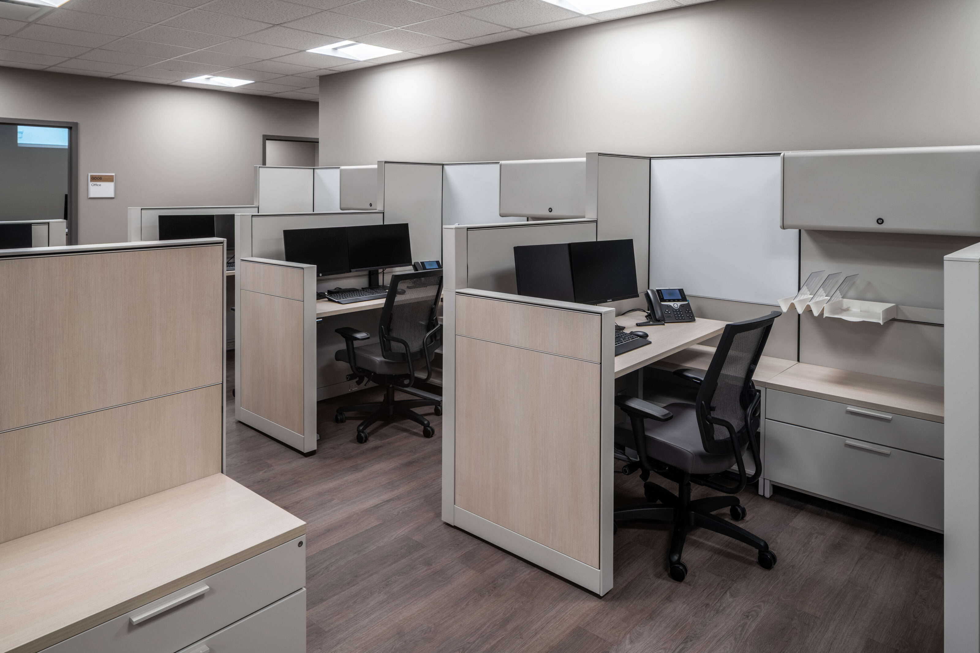 Modern office cubicles with desks, monitors, storage cabinets, and mesh chairs on wood flooring.