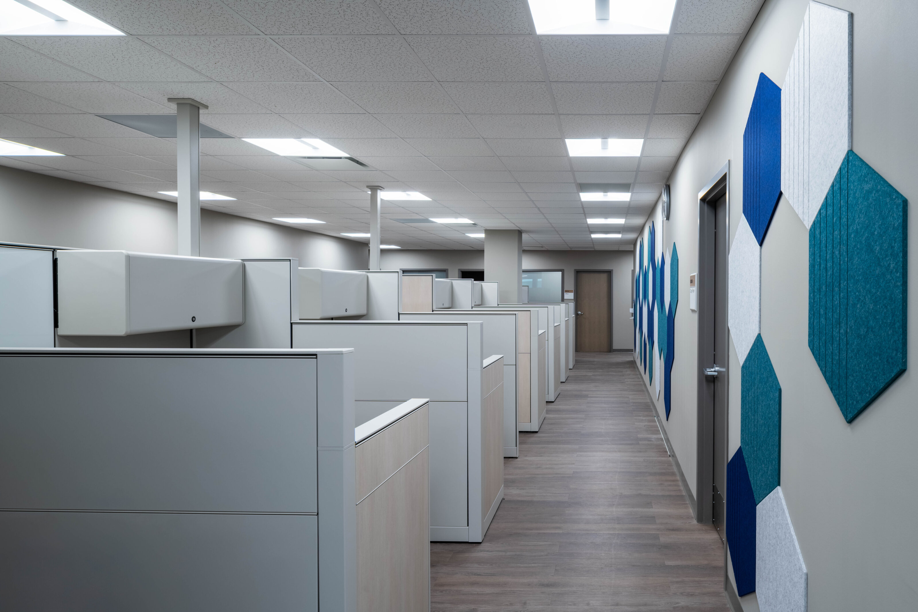 Row of office cubicles in a hallway with blue geometric wall panels and overhead lighting.