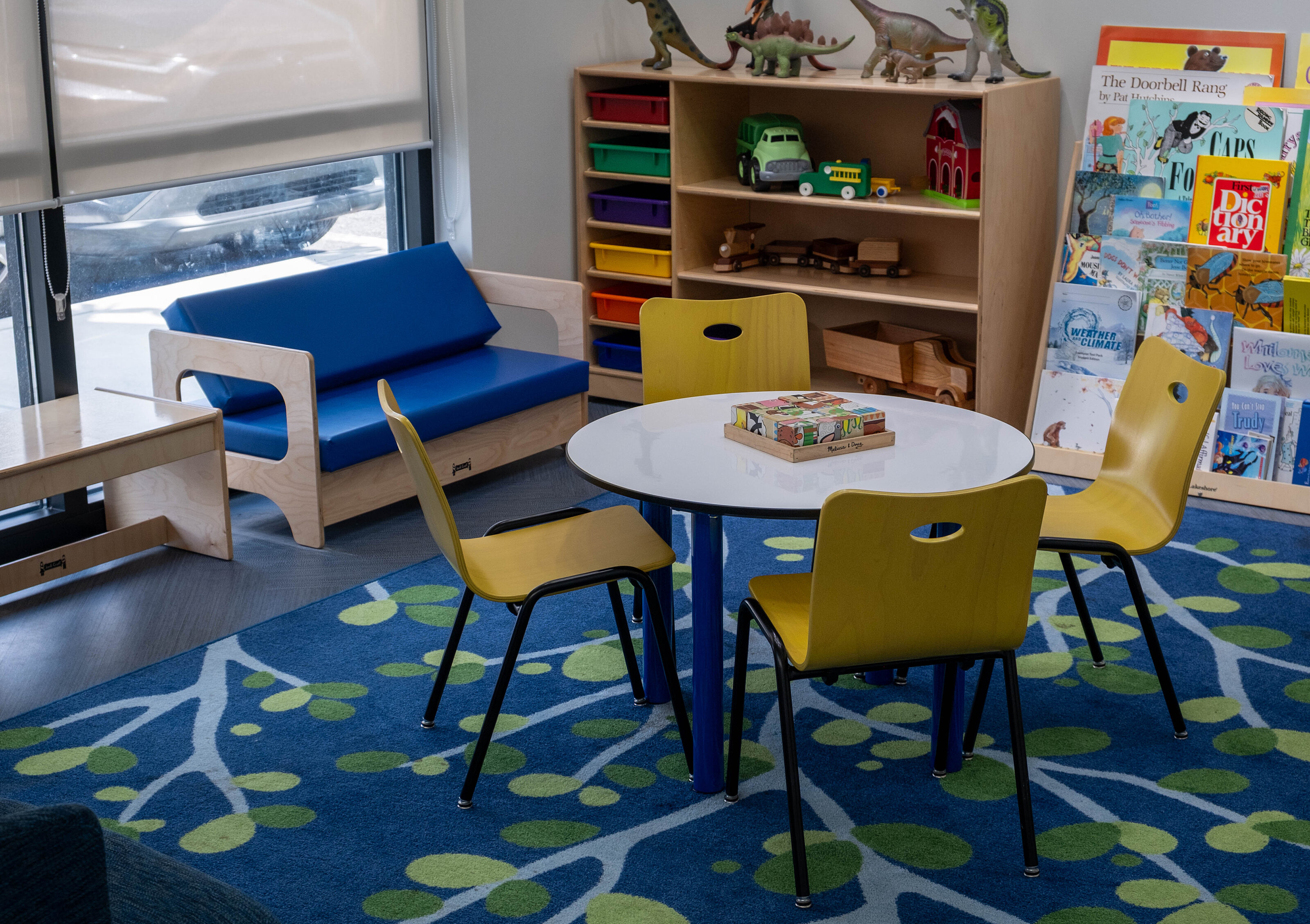 Children’s play area with round table, yellow chairs, blue sofa, shelves, and a patterned rug.