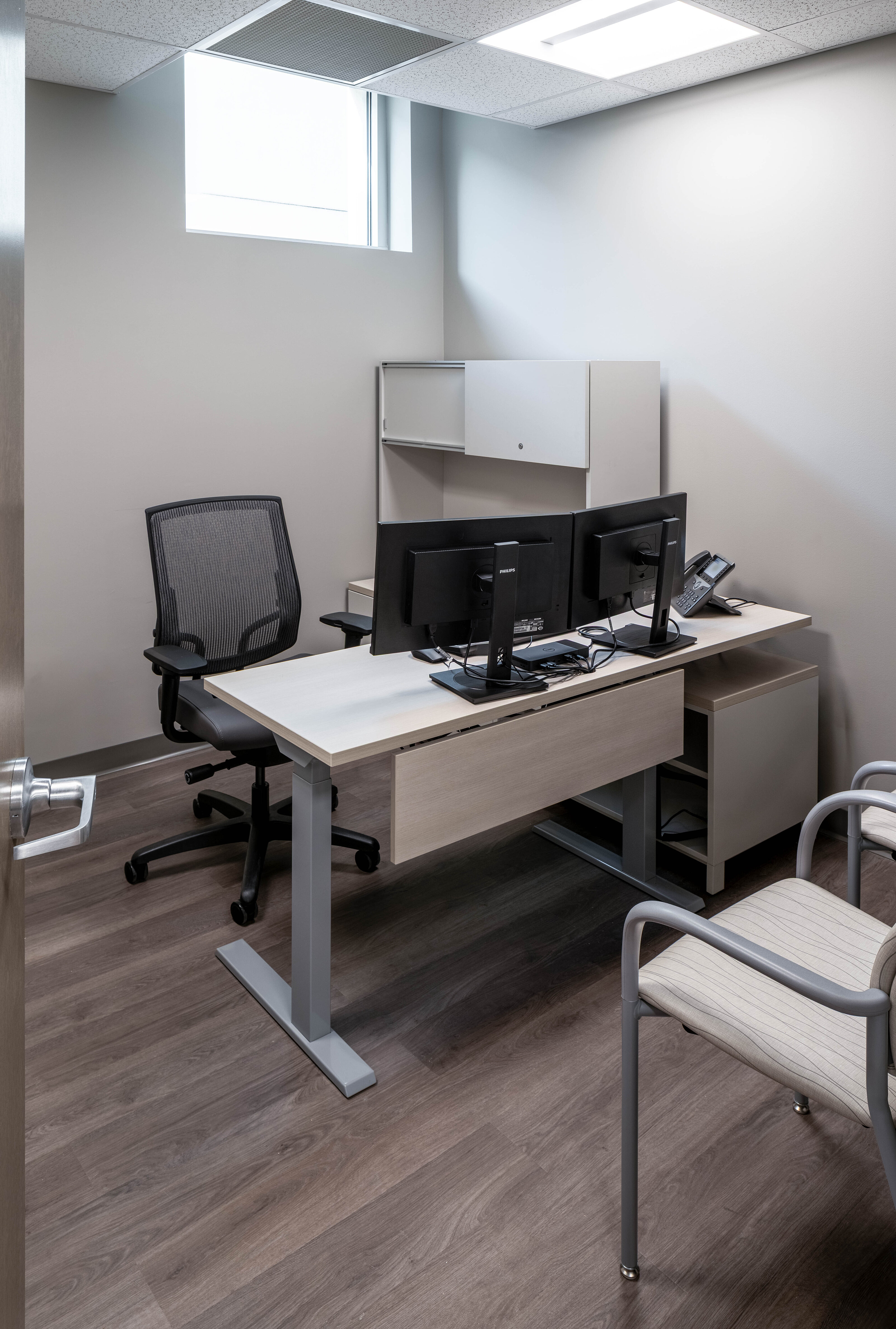 Small office with a light wood desk, dual monitors, and two chairs on a dark wood floor.