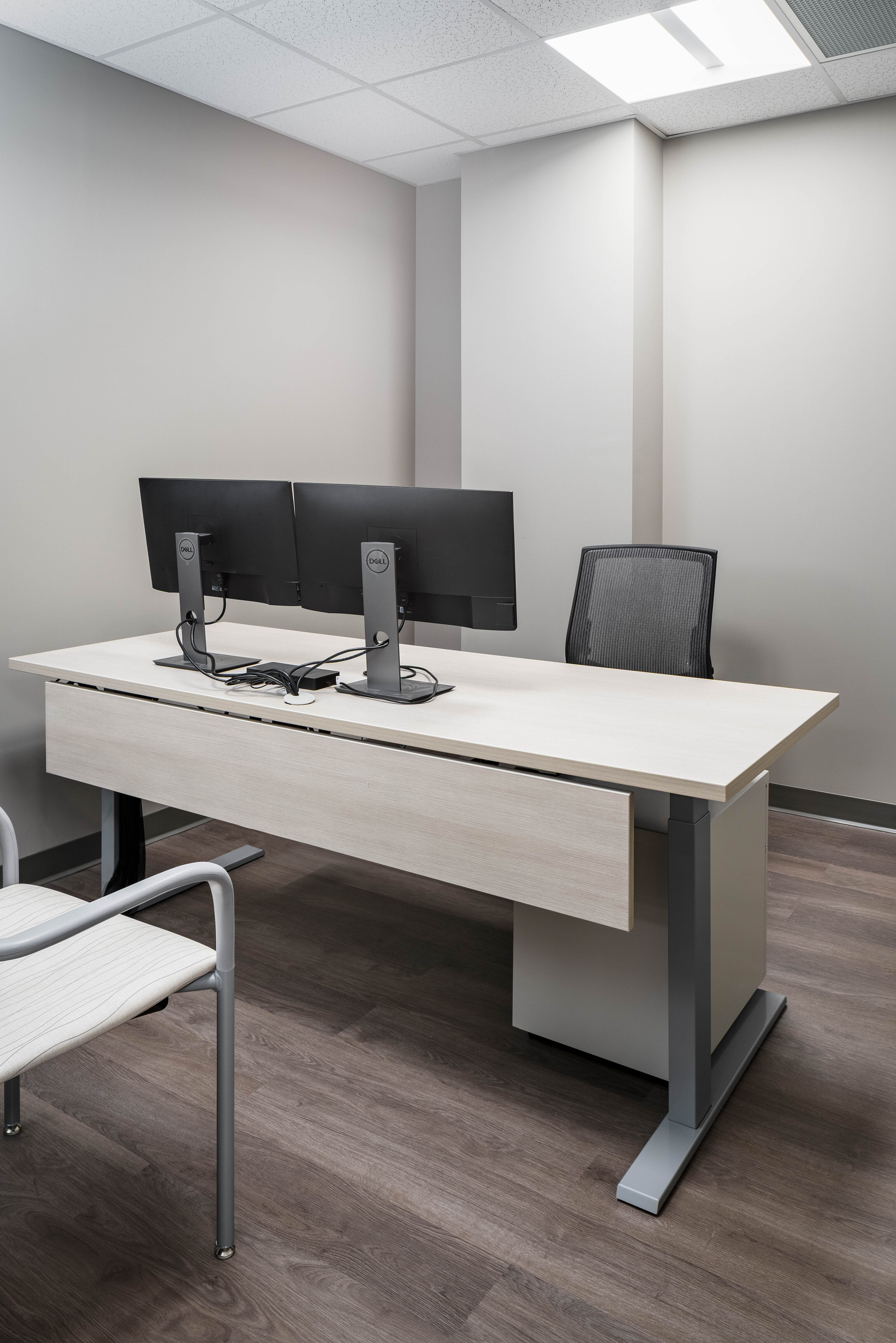 Clean office with a light wood desk, dual monitors, and a mesh chair on a dark wood floor.