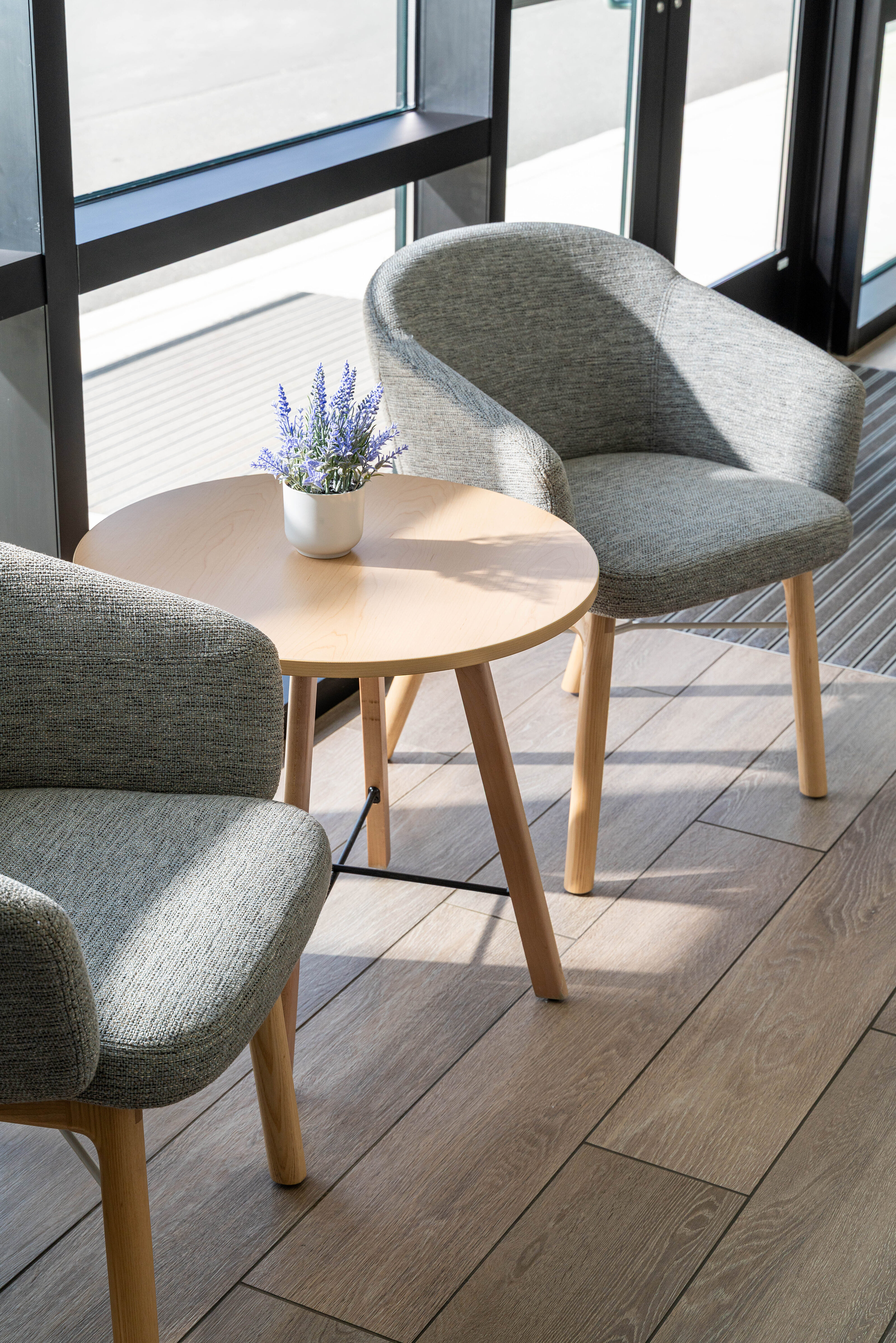 Small seating area with two gray chairs, round wood table, and potted plant by large windows.