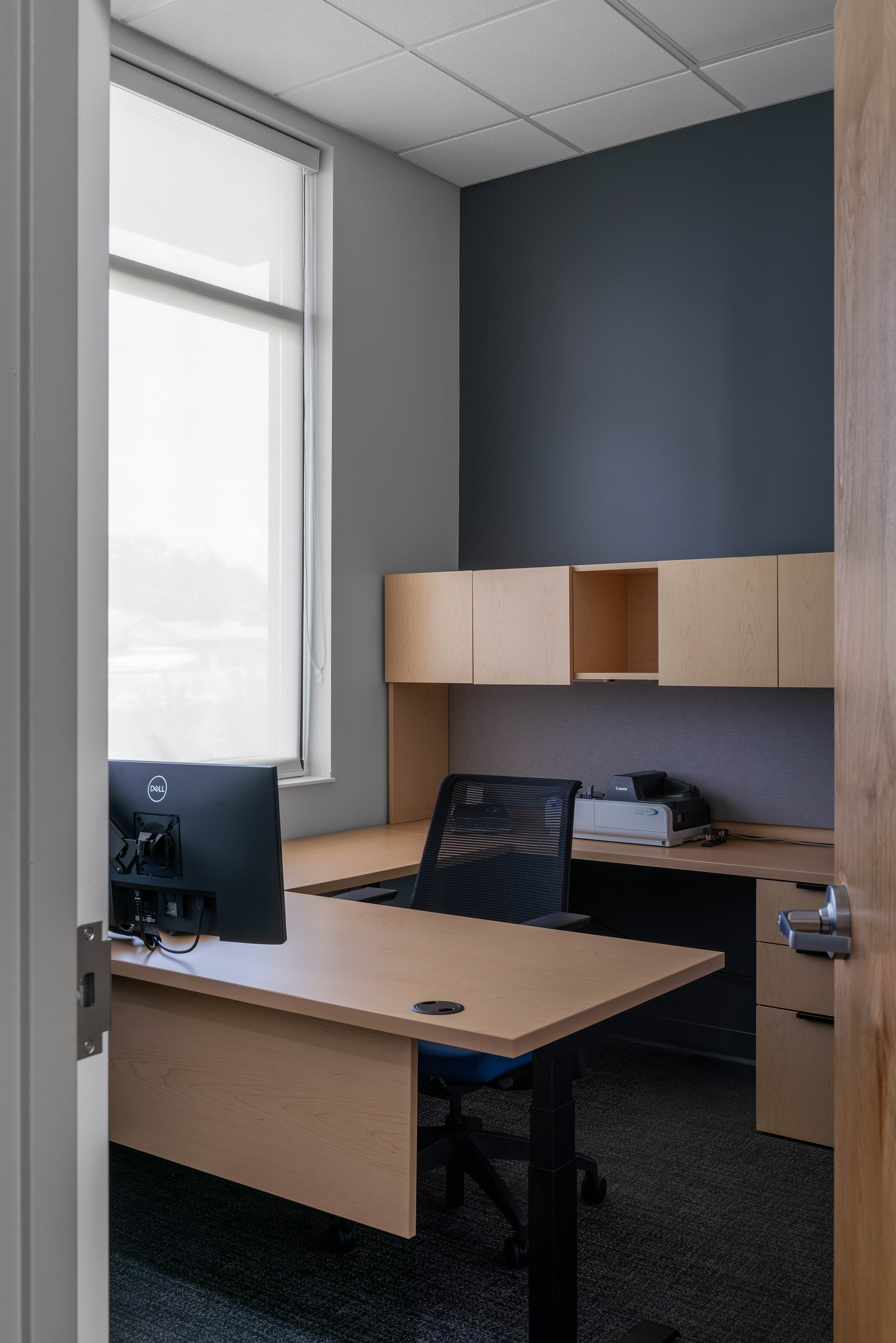 Small private office with wood desk, black chair, wall cabinets, and a window with shade.