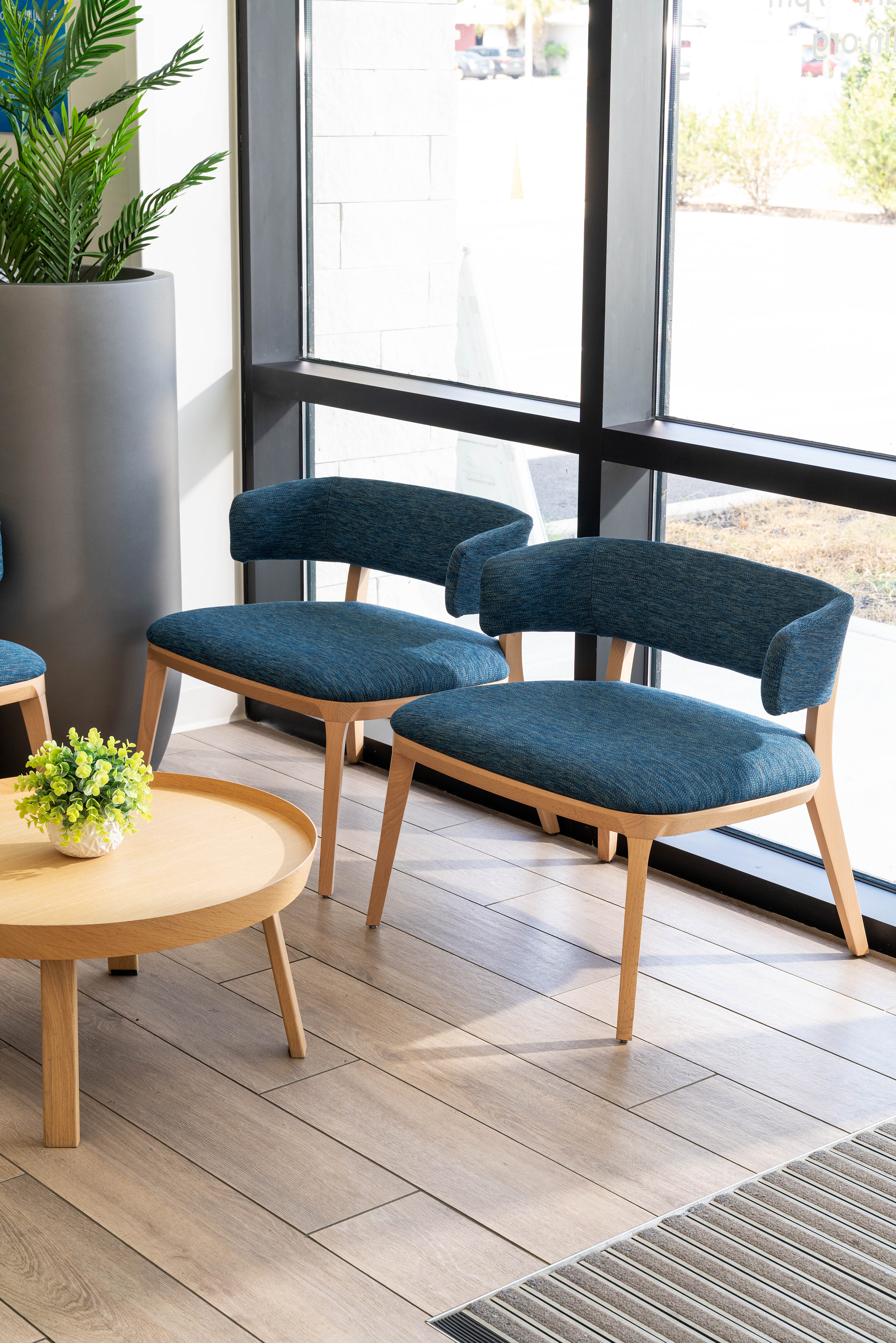 Modern waiting area with blue upholstered chairs, round wood table, and floor-to-ceiling windows.