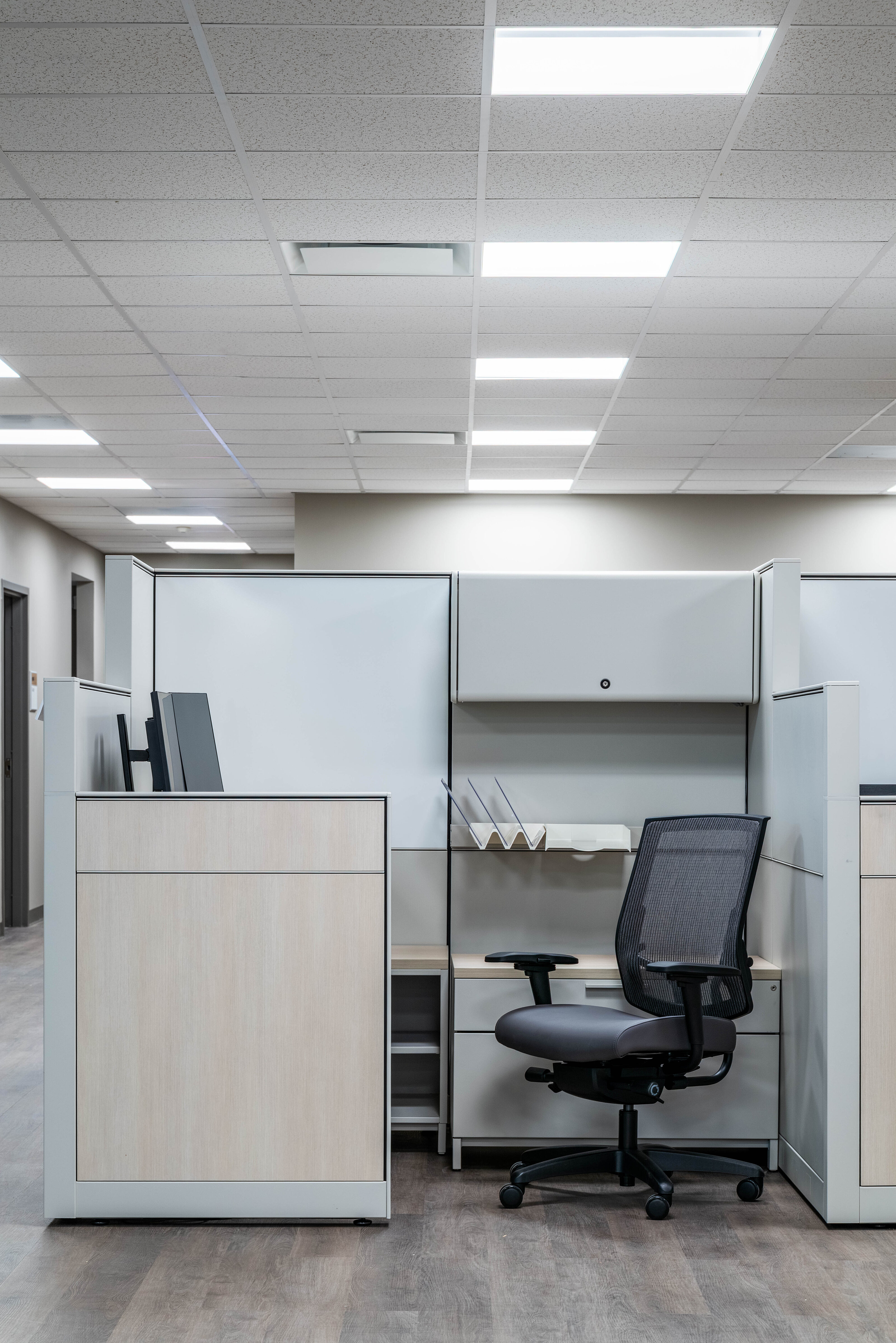 Office cubicle with a mesh chair, overhead storage, and a monitor under bright ceiling lights.