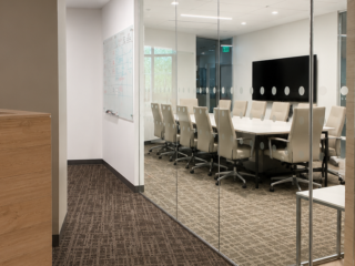 Glass-enclosed conference room featuring a meeting table with task chairs, a wall-mounted display, adjacent whiteboard, and wood-accented finishes.
