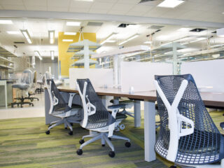 Modern open office workspace with benching desks, ergonomic task chairs, and glass lab area beyond.