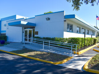 Single-story white building with a ramp entrance, blue trim, hedges, and a U.S. flag outside.