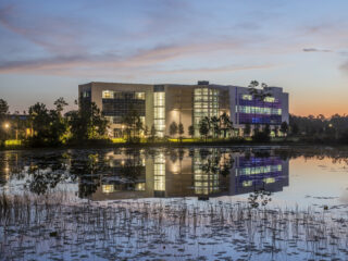 Modern campus building at dusk reflected in a calm pond with trees and sky in view.