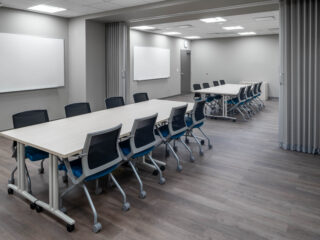 Two connected meeting rooms with long tables, rolling chairs, whiteboards, and gray partition curtains.