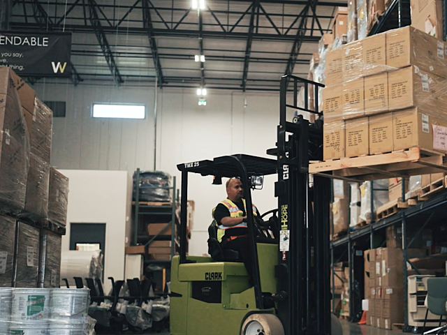 Worker operating a forklift to move stacked boxes in a warehouse aisle.