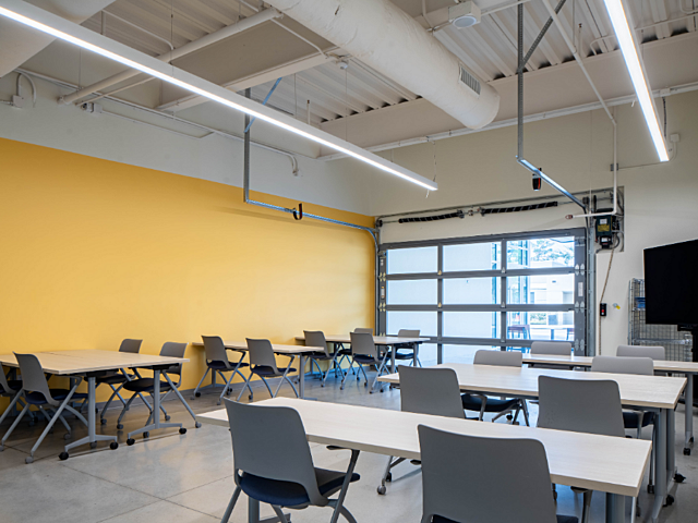 Flexible classroom with movable tables, chairs, yellow accent wall, and large glass garage door.