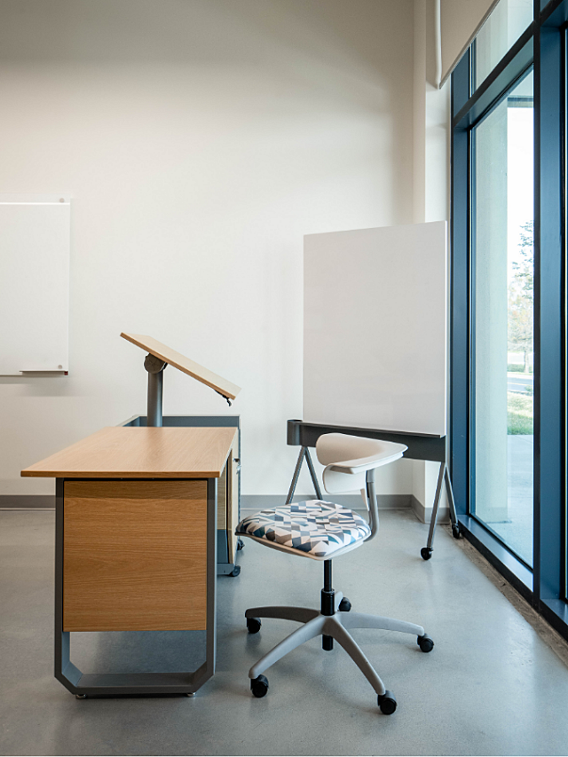 Teacher workstation with adjustable desk, rolling chair, and mobile whiteboard near windows.