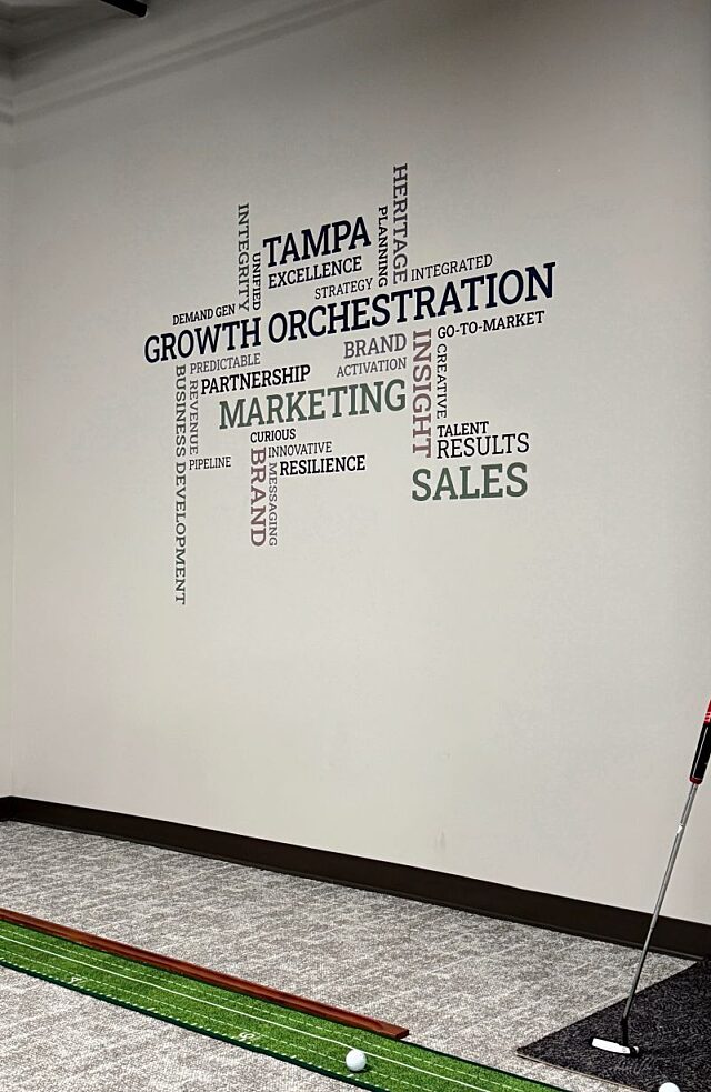 Indoor putting area with a golf mat and a wall graphic featuring Clarify’s sales and marketing values.