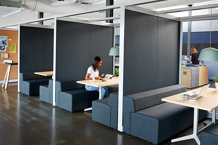 Office booth seating area with upholstered benches and tables, a person working at a laptop in one booth.
