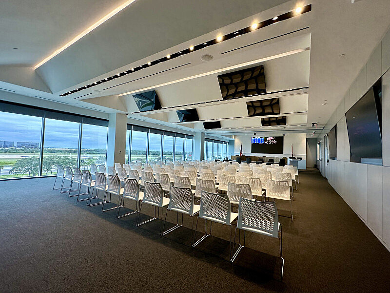 Conference room with rows of white chairs facing a presentation area, floor-to-ceiling windows along one side.