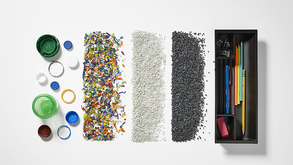 Plastic bottle caps and sorted plastic pellets arranged beside an open drawer of office supplies.
